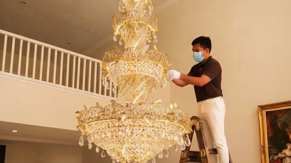 A MELLS technician on a ladder cleaning a gilt and crystal chandelier by hand in a double height Dubai hallway